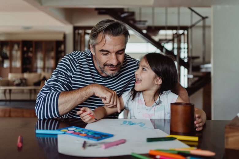 a father sitting beside his daughter pointing the blue paint on the paper